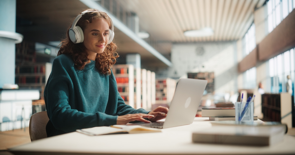 A woman in a library working on her computer with headphones on.