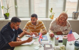 Tres adultos que estudian inglés juntos en una mesa de aula con pequeñas banderas estadounidenses y británicas.