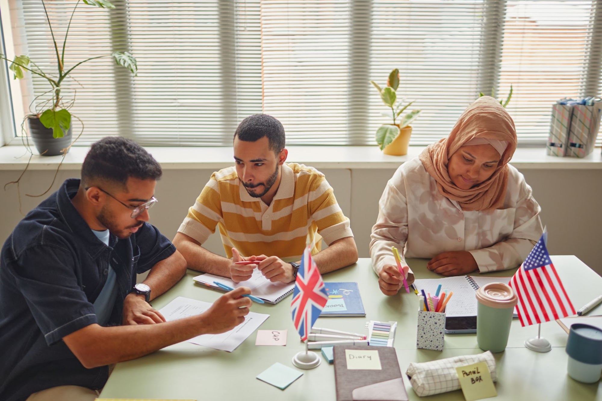Three adult learners studying English together at a classroom table with small American and British flags